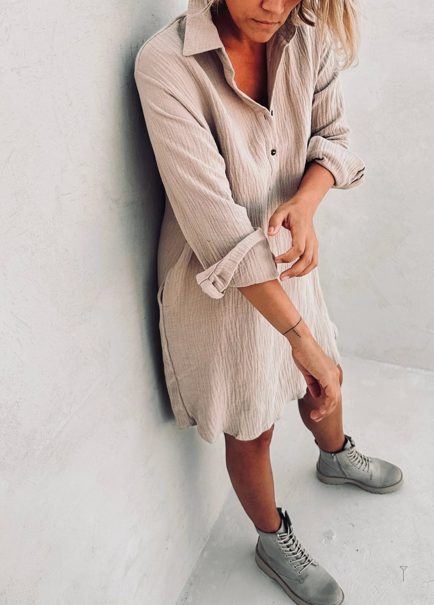 A woman in a beige shirt dress leaning against the wall.