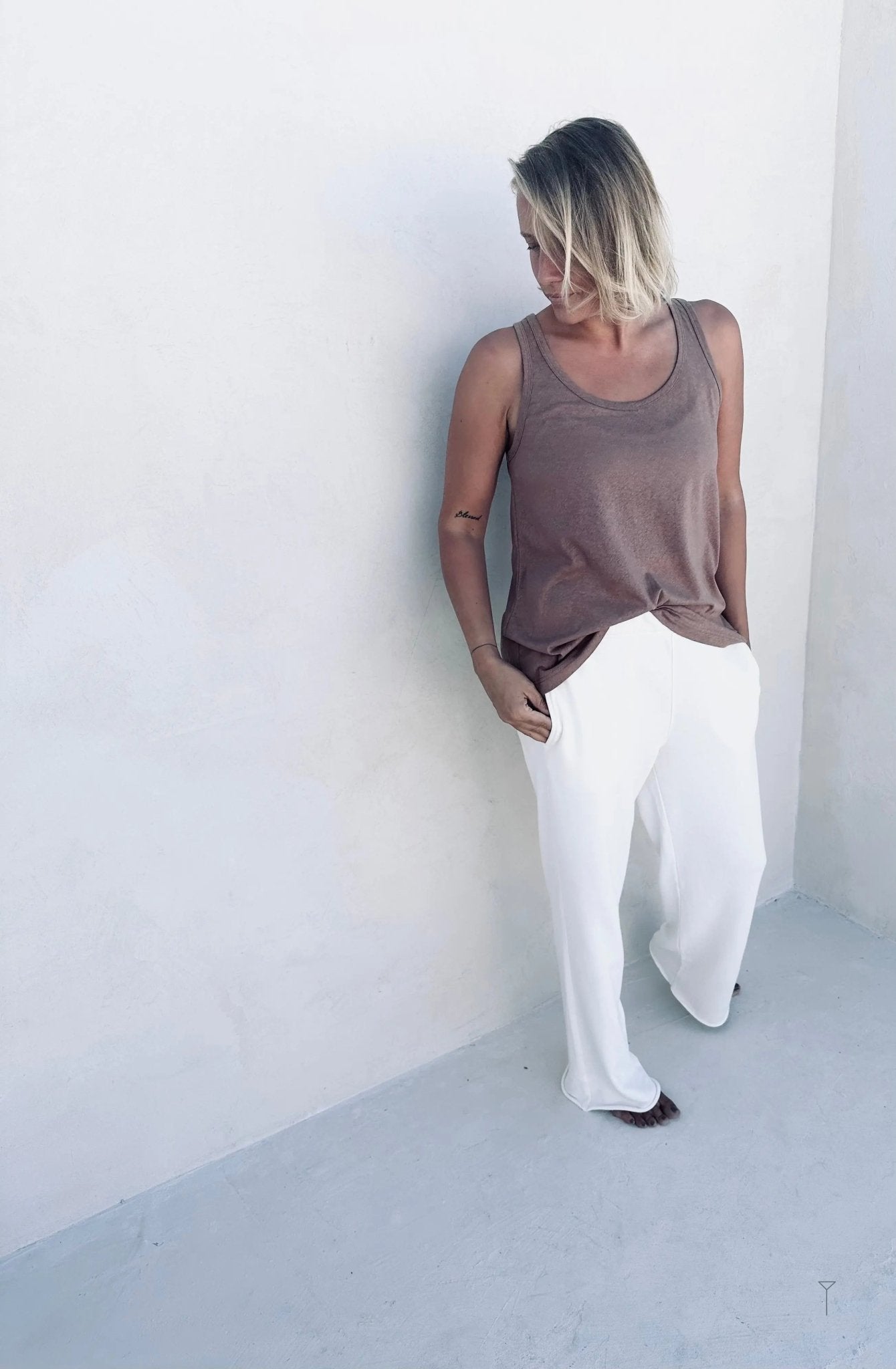 A woman leaning against a wall wearing a brown tank top and white pants.