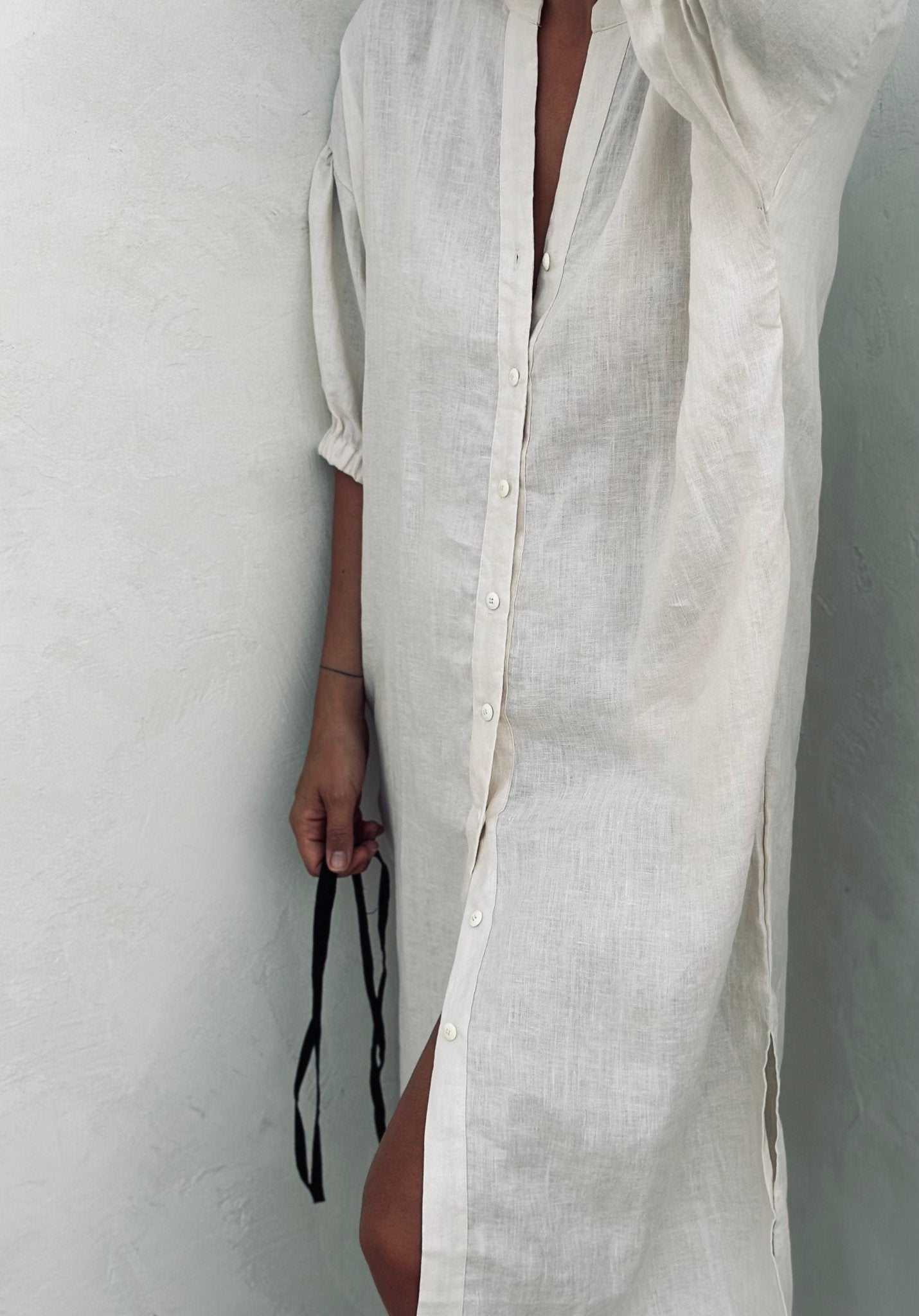 A woman in a white linen dress leaning against the wall. 